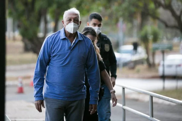foto colorida de homem de blusa azul e máscara facial cercado por policiais