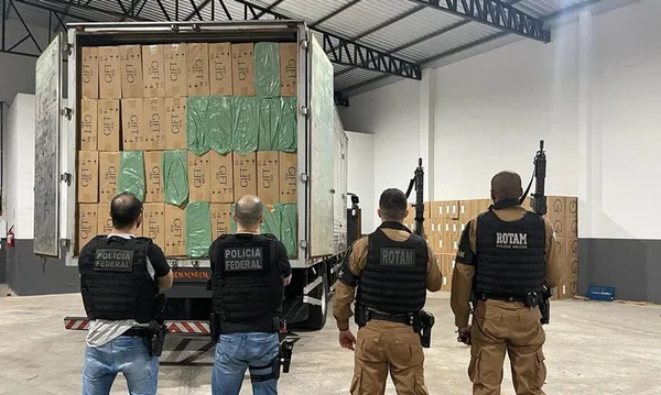 Imagem colorida de agentes da PF de costas e uma carga de cigarros contrabandeados apreendidos - Metrópoles