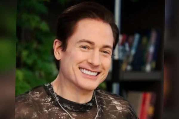 Foto colorida de homem branco, usando blusa de tule cinza, sorrindo para foto - Metrópoles.