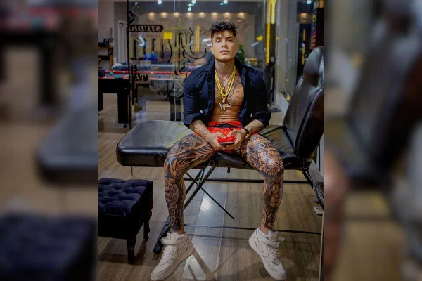 Iamgem colorida de homem coberto com tatuagens enquanto olha para câmera sentado em maca preta - Metrópoles