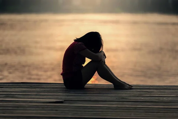 Mulher triste em sentada com a cabeça abaixada sob os joelhos. Ela está num deck e ao fundo é possível ver um lago sob o entardecer - Metrópoles