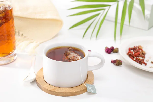 Na foto temos uma xícara branca com um liquido escuro e ums ache de cha dentro e ao redor um vidro com liquido em tom caramelo, um prato com sementes e a ponta de uma folha de palmeira