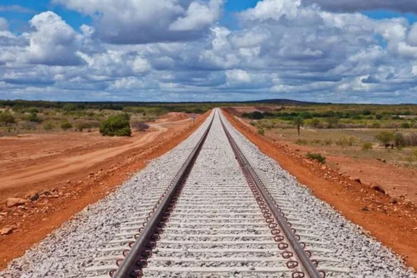 Imagem de trilho de ferrovia cortando uma área de terra e vegetação, com o horizonte ao fundo e céu azul com nuvens - Metrópoles