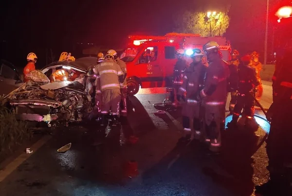 Bombeiros, à noite, atendendo a vítima de capotamento. Carro prata aparece destruído