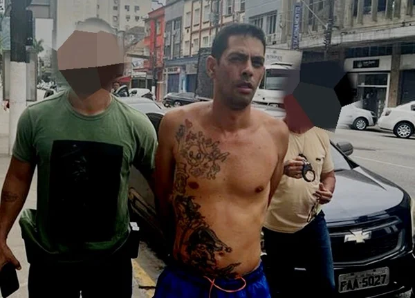 Em foto colorira homem sem camisa, tatuagens na lateral do corpo, é conduzido algemado, com mas mãos para trás, por dois policiais - Metrópoles