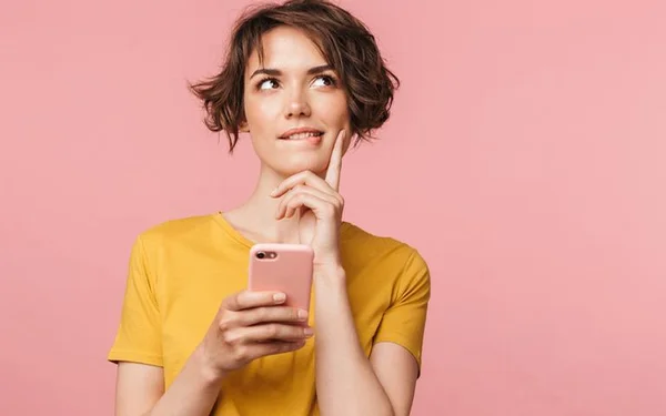 Mulher com celular na mão com uma blusa amarela em um fundo rosa