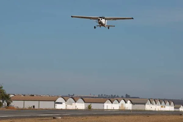 Fotografia colorida de avião chegando em pista de pouso com hangares ao fuundo