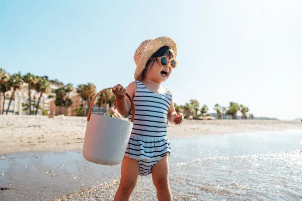 Imagem mostra uma criança pequena, de maiô listrado, óculos escuros e balde na mão. Ela está se divertindo na praia - Metrópoles