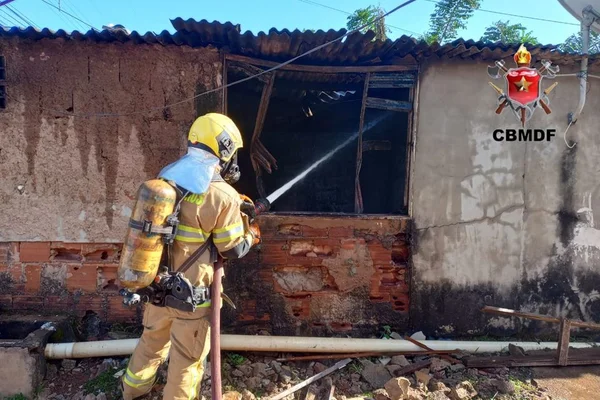 Homem com uniforme de bombeiro de costas segurando mangueira com água e apontando para janela de casa